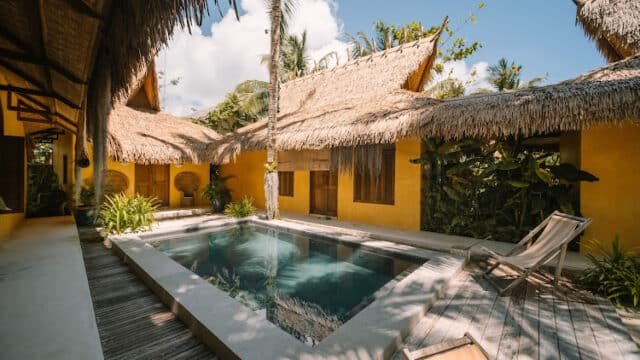 A tropical Siargao courtyard with a small pool, lounge chairs, and yellow thatched-roof buildings under a blue sky.