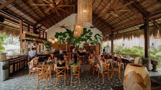 Open-air Siargao restaurant with wooden decor, leafy plants, and people dining at tables under a thatched roof.