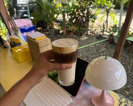 A hand holds an iced coffee above a desk with a tablet, keyboard, and lamp in a Siargao outdoor garden cafe.