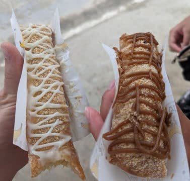 Two hands holding churros, one with white icing and the other with caramel drizzle, enjoyed on a Siargao-inspired concrete background.