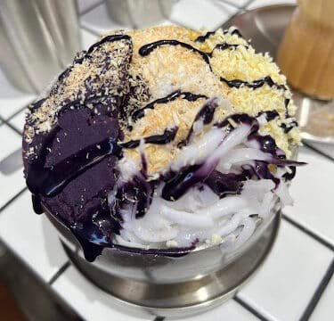 A bowl of halo-halo topped with ube, shredded coconut, flan, and sweet syrup on a tiled table in sunny Siargao.
