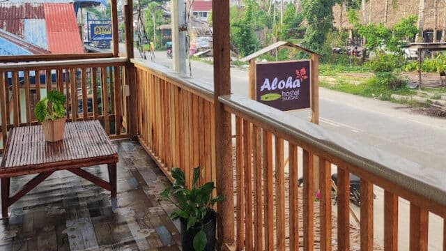 A bamboo balcony with potted plants overlooks a Siargao street and a sign that reads Aloha Hostel Siargao.