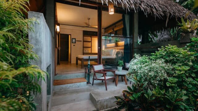 Tropical Siargao bungalow with a thatched roof, patio chairs, and lush greenery outside a cozy bedroom.