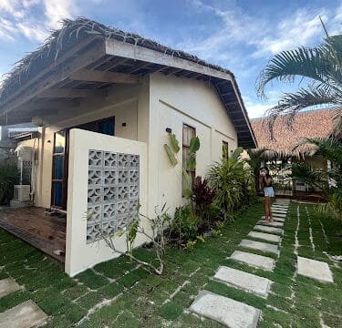 A small white bungalow with a thatched roof in Siargao, surrounded by greenery and a paved stone walkway under a blue sky.