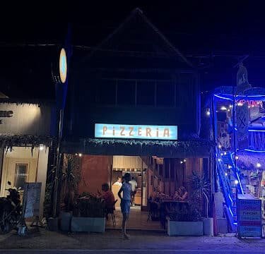A person stands outside a lit-up pizzeria in Siargao at night, with people dining inside and neon lights glowing nearby.