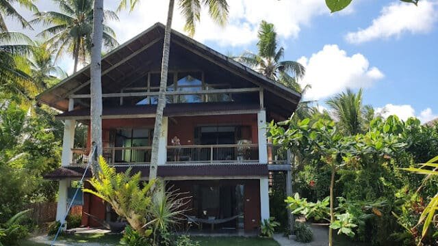 A tropical two-story Siargao house surrounded by palm trees and lush green garden under a sunny sky.