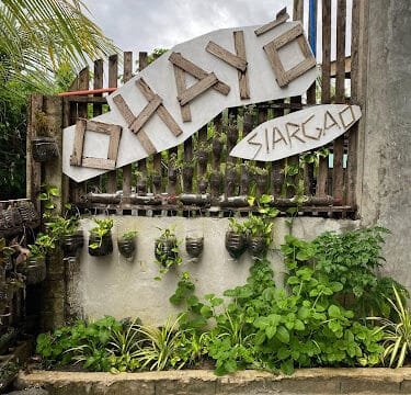Wooden sign reading Ohayo Siargao on a fence, surrounded by lush Siargao greenery and vibrant plants below.