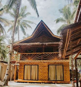 A two-story bamboo hut with large windows, nestled among palm trees on sandy Siargao ground.
