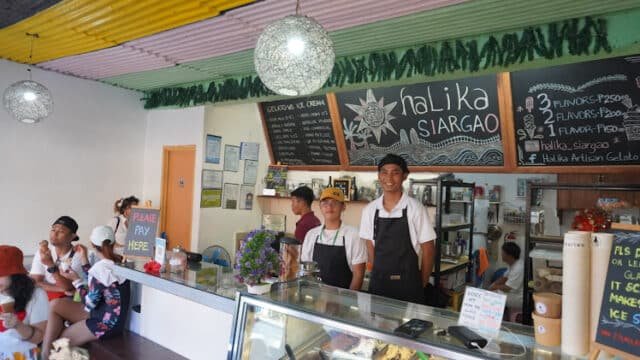 Two staff in aprons stand behind the counter of Halika Siargao gelato shop, with customers seated inside on a sunny Siargao afternoon.