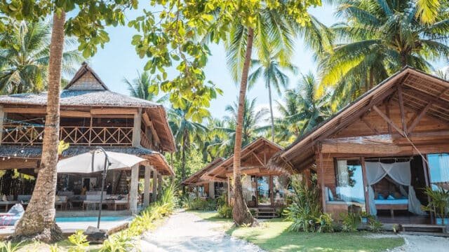Tropical Siargao beach resort with wooden huts, palm trees, and a sandy path under bright, sunny weather.