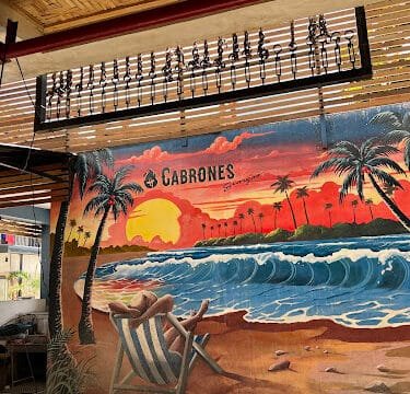 Mural of a Siargao beach sunset with palm trees, waves, and a person relaxing in a chair under a sign that says “CABRONES.”