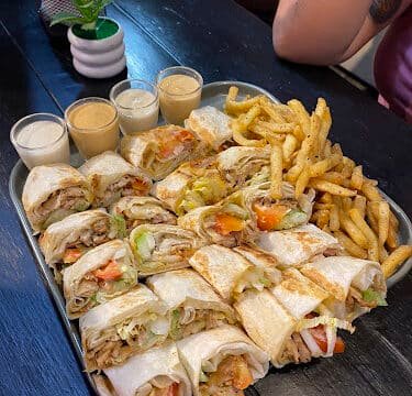 A tray of wraps, fries, and dipping sauces on a dark table in Siargao, with drinks and potted plants in the background.