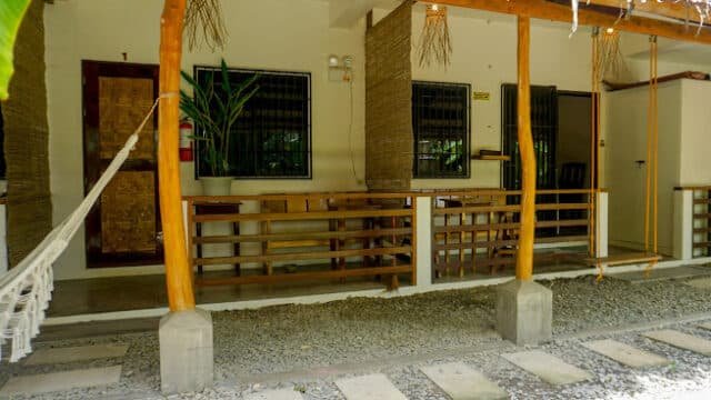 A porch in Siargao with wooden railings, a hammock, a swing, and a bench outside a building with two doors and windows.