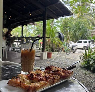 Iced coffee and pizza on a table at an outdoor café in Siargao, surrounded by greenery and parked bicycles in the background.