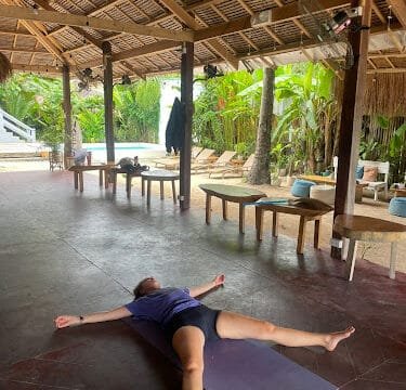 Person lying on a yoga mat in a relaxed pose under an open-air patio with tropical plants and a Siargao pool nearby.