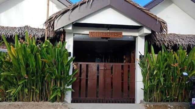 Front view of a small Siargao building with thatched roof and tall green plants by the entrance gate.