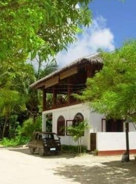A white two-story house with a thatched roof in Siargao, surrounded by green trees and a parked black car.