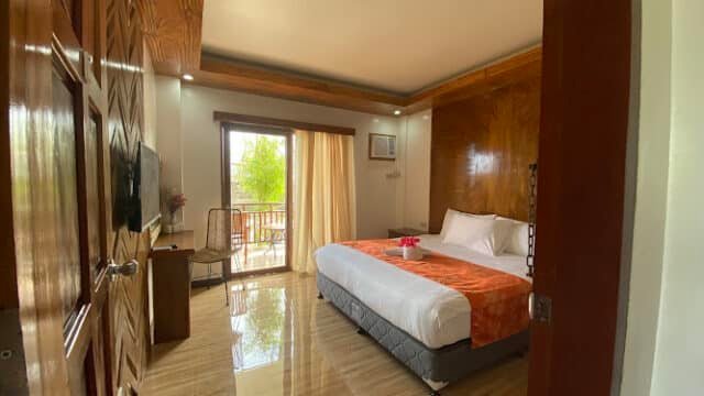 A bright Siargao hotel room with a king bed, desk, TV, balcony, and wooden accents seen from the doorway.