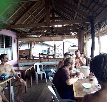 People sitting and eating at wooden tables in an open-air, thatched-roof Siargao restaurant or café.