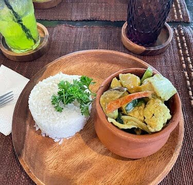 A wooden plate with white rice and vegetable curry, served Siargao-style with a green drink on a woven placemat.