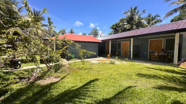 Sunny yard with green grass, trees, and two small buildings with brown doors under Siargao’s clear blue sky.
