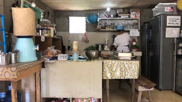 A person prepares food in a small, rustic Siargao kitchen with shelves, a fridge, and various utensils visible.