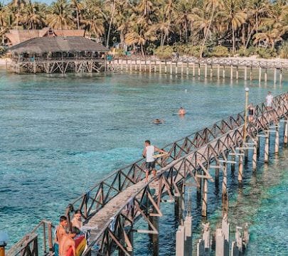 Wooden pier over clear blue water in Siargao, people walking, and palm trees lining the shore in the background.