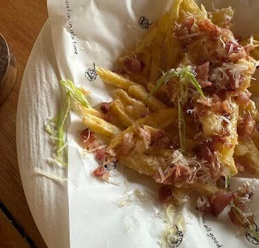 French fries topped with grated cheese, bacon bits, and green onions, served Siargao-style on white paper in a dish.