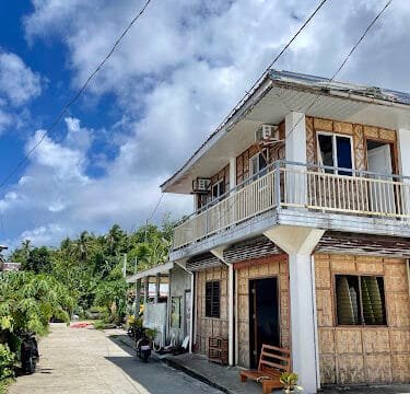 A two-story house in Siargao with a balcony, surrounded by lush plants under a clear blue sky with clouds on a sunny day.