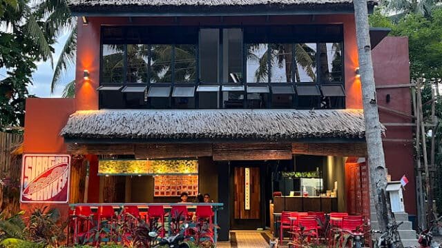 A two-story tropical Siargao restaurant with red chairs, thatched roof, and palm trees, seen from the front at dusk.