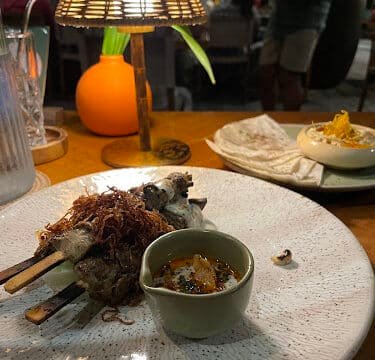 Three skewers with sauce on a plate, set on a Siargao restaurant table with a lamp and other dishes in the background.