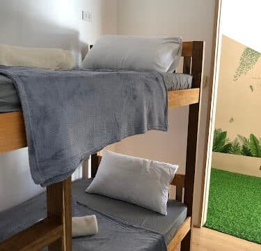 Wooden bunk beds with gray bedding beside an open door in Siargao, leading to a sunlit area with green artificial grass.
