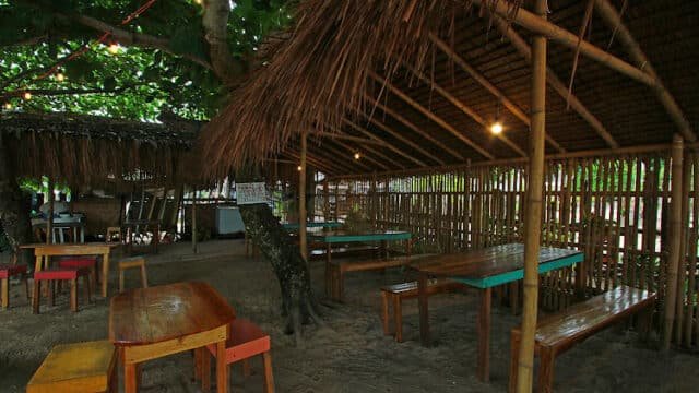 Outdoor bamboo hut restaurant in Siargao with wooden tables, benches, and sandy floor under leafy trees, softly lit by bulbs.