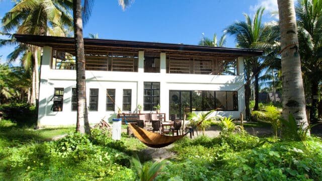 A modern two-story house in Siargao, surrounded by palm trees, outdoor furniture, and a hammock in a lush green yard.