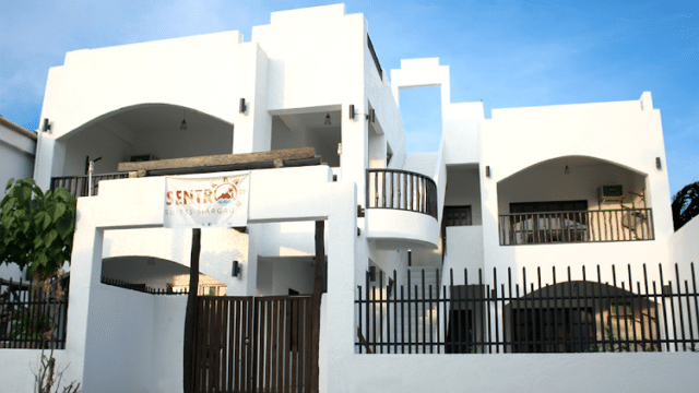 White two-story Siargao building with balconies and arched doorways, behind a dark fence, under a blue sky.