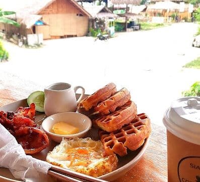 Plate with waffles, fried egg, bacon, and syrup, plus coffee, enjoyed with a stunning Siargao village view in the background.
