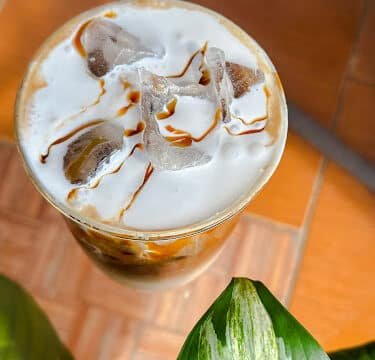 Iced coffee with cream and syrup, topped with ice cubes, sits next to a green leaf on a tiled Siargao-inspired surface.