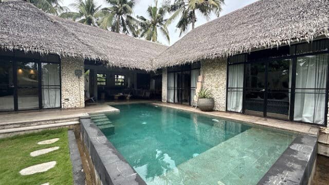 A Siargao villa with a thatched roof, private pool, stone walls, and palm trees in the background.