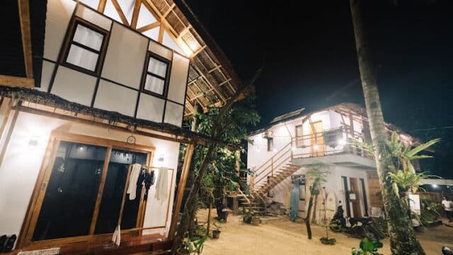 Two white, wooden cottages with balconies are lit up at night, surrounded by tropical plants and Siargao’s sandy ground.