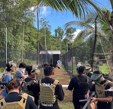 People in tactical vests gather outdoors in Siargao, aiming at targets on a fenced paintball or airsoft field under blue sky.