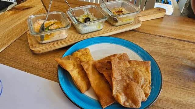 Rectangular pita chips on a blue plate with three dips in glass bowls, inspired by the flavors of Siargao, on a wooden board behind.