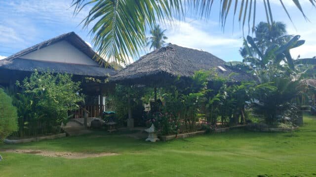 Thatched-roof huts in Siargao surrounded by lush tropical plants and green grass under a partly cloudy sky.