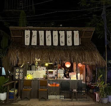 A small, thatched-roof cafe in Siargao at night, glowing with warm lights and handwritten signs above the entrance.