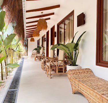 Wicker chairs and plants line a sunlit outdoor patio with wooden beams and large windows, evoking a laid-back Siargao vibe.