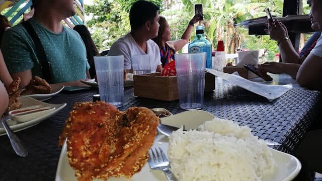 A plate of fried chicken and rice on a table in Siargao, with people dining and taking photos outdoors.