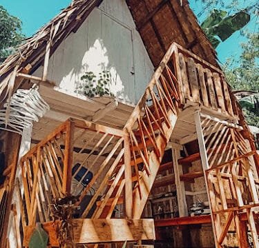 A-frame wooden cabin in Siargao with an outdoor staircase, surrounded by greenery under a clear blue sky.