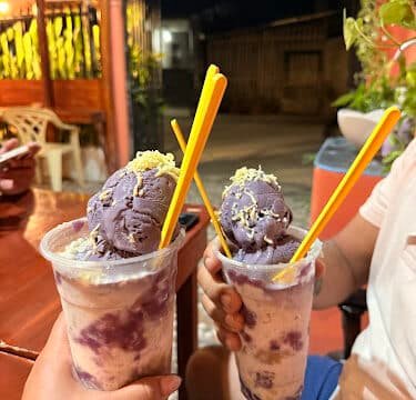 Two hands holding cups of halo-halo with purple ice cream and cheese, enjoyed outdoors on a warm Siargao night.