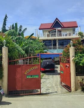 A two-story house in Siargao with a red roof, a red gate labeled “NOGUERRA WELCOME,” and a car parked inside.
