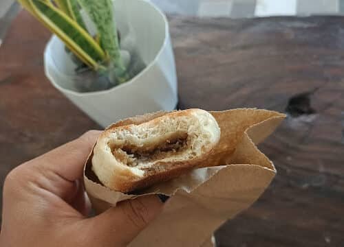 A hand holds a half-eaten bread roll in a brown paper bag, with a potted plant and Siargao vibes in the background.