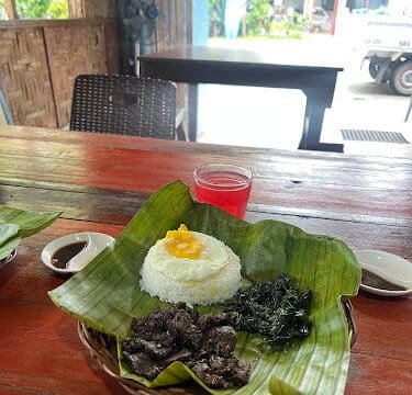 A Siargao meal with rice, beef, greens on banana leaves, and a red drink on a wooden table in a rustic setting.